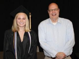 2 people standing in front of black backdrop with one wearing graduation cap & gown